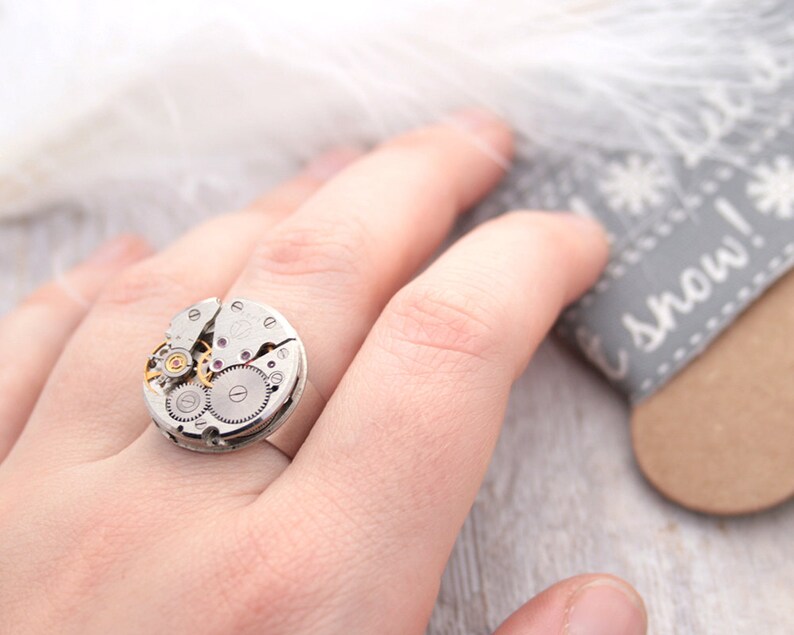 May include: A silver ring with a circular watch face design. The watch face is made of metal and has visible gears and a small pink jewel.