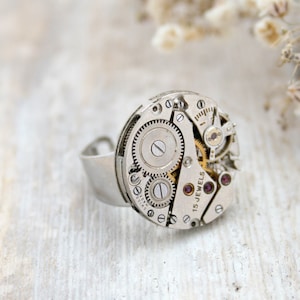 Peut inclure: Une bague en argent avec un cadran de montre comme pièce maîtresse. Le cadran de montre a des engrenages apparents et le texte "15 JEWELS".