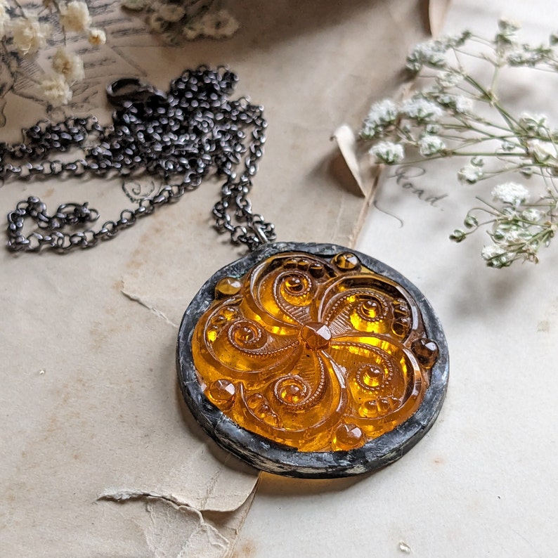Puede incluir: Un collar con un colgante de vidrio dorado y &aacute;mbar con un dise&ntilde;o floral detallado. El colgante est&aacute; engastado en un marco de metal plateado y cuelga de una cadena plateada.