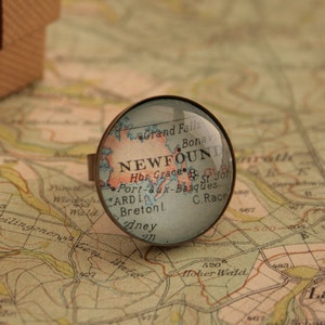May include: A vintage-style glass ring featuring a map of Newfoundland, Canada. The map includes the text "NEWFOUND" and "C. Race".