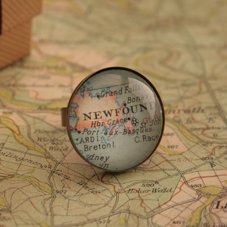 May include: A vintage-style glass ring featuring a map of Newfoundland, Canada. The map includes the text "NEWFOUND" and "C. Race".