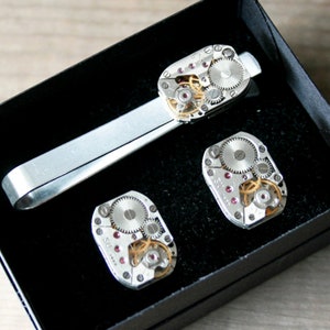 Könnte beinhalten: Ein Set silberner Manschettenknöpfe und einer Krawattenklammer mit filigranen Uhrwerkdesigns. Die Manschettenknöpfe und die Krawattenklammer sind aus Metall und haben eine silberne Oberfläche. Die Uhrwerkdesigns sind durch das klare Glas sichtbar.