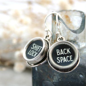 May include: Silver earrings featuring typewriter keys. The keys are black with white lettering. One key reads "SHIFT LOCK" and the other reads "BACK SPACE".