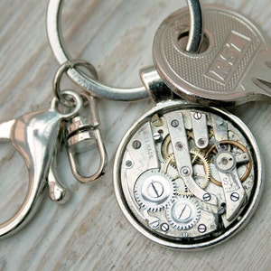 May include: A silver keychain featuring a round pendant showcasing the intricate internal gears of a watch. The pendant is attached to a key ring and a clasp. A key with the text "RST" is also present.