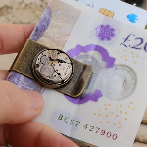 May include: A vintage-style money clip with a watch mechanism centerpiece. The clip is made of aged brass and holds a twenty-pound note. The watch mechanism features visible gears and intricate details, adding a unique touch to the accessory.