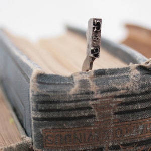 May include: Close-up of a vintage book with a metal bookmark. The bookmark is shaped like a small, rectangular piece of metal with the letters "G" and "5" engraved on it. The book is bound in brown leather and has a worn, aged appearance.