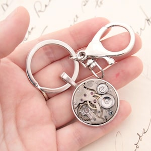May include: Silver key chain with a round watch face charm. The watch face is open and shows the inner workings of the watch.