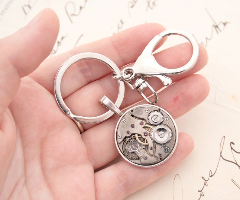 May include: Silver key chain with a round watch face charm. The watch face is open and shows the inner workings of the watch.