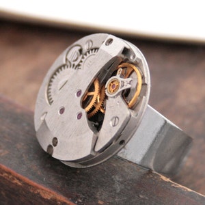 May include: A silver ring with a watch face embedded in the top. The watch face is open and shows the inner workings of the watch, including gears and a red jewel.