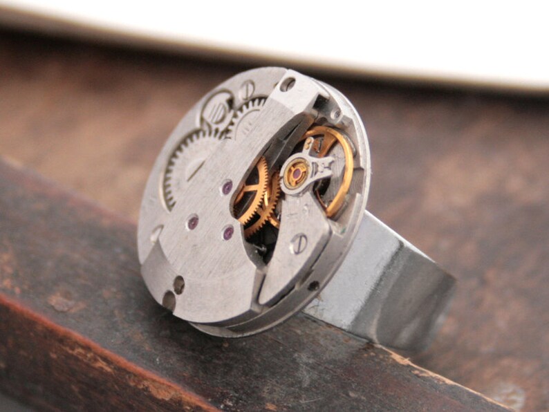 May include: A silver ring with a watch face embedded in the top. The watch face is open and shows the inner workings of the watch, including gears and a red jewel.