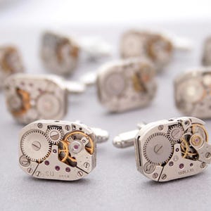 May include: Silver cufflinks with exposed watch mechanisms. The rectangular cufflinks display intricate gears, springs, and jewels, showcasing a detailed mechanical design. The cufflinks have a polished finish, offering a classic, elegant style.