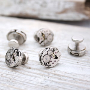May include: Set of four silver cufflinks featuring intricate watch gears and a small pink jewel. The cufflinks are arranged on a light wood surface.
