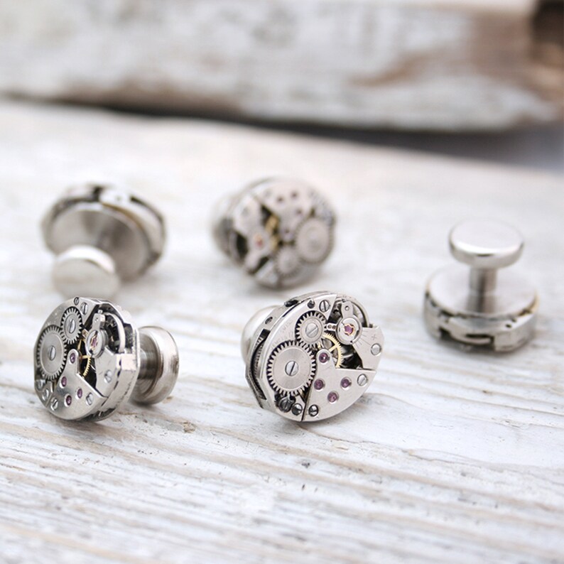 May include: Set of four silver cufflinks featuring intricate watch gears and a small pink jewel. The cufflinks are arranged on a light wood surface.