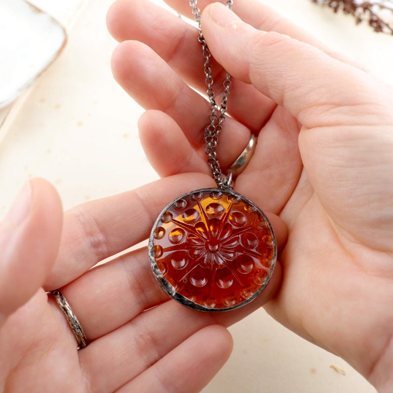 Puede incluir: Un collar de cadena plateada con un colgante redondo de vidrio naranja con un dise&ntilde;o floral en relieve detallado.