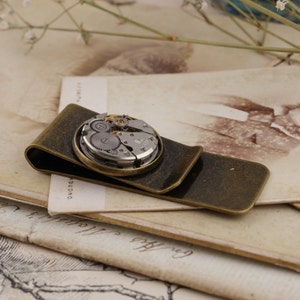 May include: A vintage-style money clip with a watch mechanism centerpiece. The clip is made of bronze-toned metal and features a circular watch movement with visible gears and intricate details. The clip is resting on a stack of aged paper.