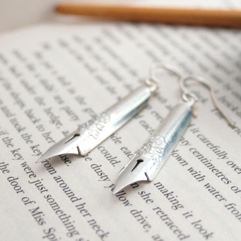 Puede incluir: Pendientes de plata con forma de plum&iacute;n. Los pendientes est&aacute;n sobre un libro con texto.