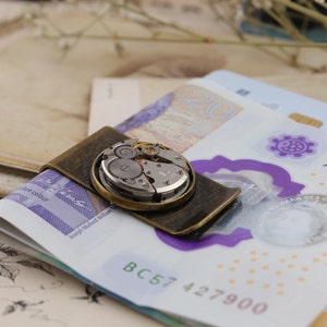 May include: A vintage-style money clip with an antique brass finish, holding several bills. The clip features a circular inset showcasing the intricate gears and mechanisms of a watch movement. The bills are visible beneath the clip.