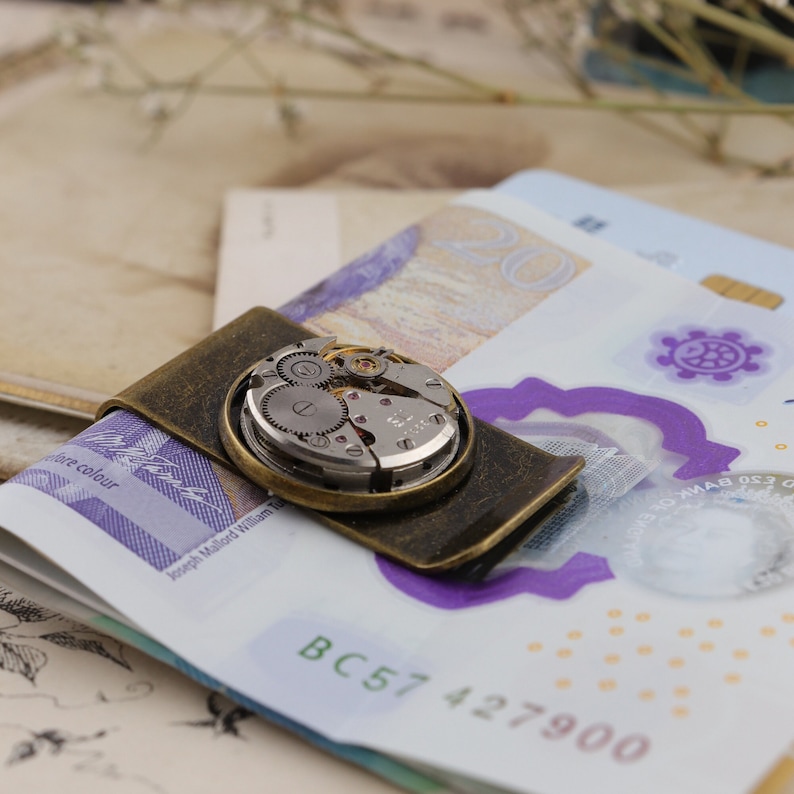 May include: A vintage-style money clip with an antique brass finish, holding several bills. The clip features a circular inset showcasing the intricate gears and mechanisms of a watch movement. The bills are visible beneath the clip.