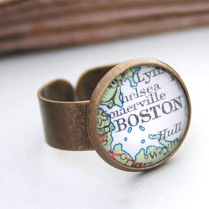 Può includere: Un anello in ottone con un cabochon rotondo in vetro che presenta una mappa vintage di Boston, Massachusetts. La mappa mostra i nomi dei quartieri, tra cui Chelsea, Somerville e Hull.