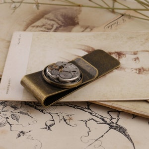 May include: A vintage-style money clip in a bronze finish, featuring a circular watch mechanism as a decorative element. The watch parts are visible within the circle. The clip is resting on a vintage postcard.