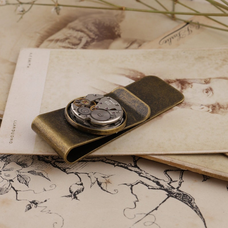 May include: A vintage-style money clip in a bronze finish, featuring a circular watch mechanism as a decorative element. The watch parts are visible within the circle. The clip is resting on a vintage postcard.