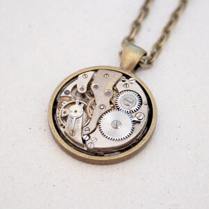 May include: A bronze necklace with a round pendant featuring a visible clockwork mechanism. The pendant is made of metal and has a circular shape. The clockwork is visible through a clear glass or plastic cover.