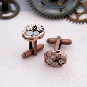 May include: A pair of copper-toned cufflinks featuring exposed watch gears and mechanisms. The oval-shaped faces showcase intricate details of the internal workings, with small gears and other components visible. The cufflinks have a classic, elegant design.