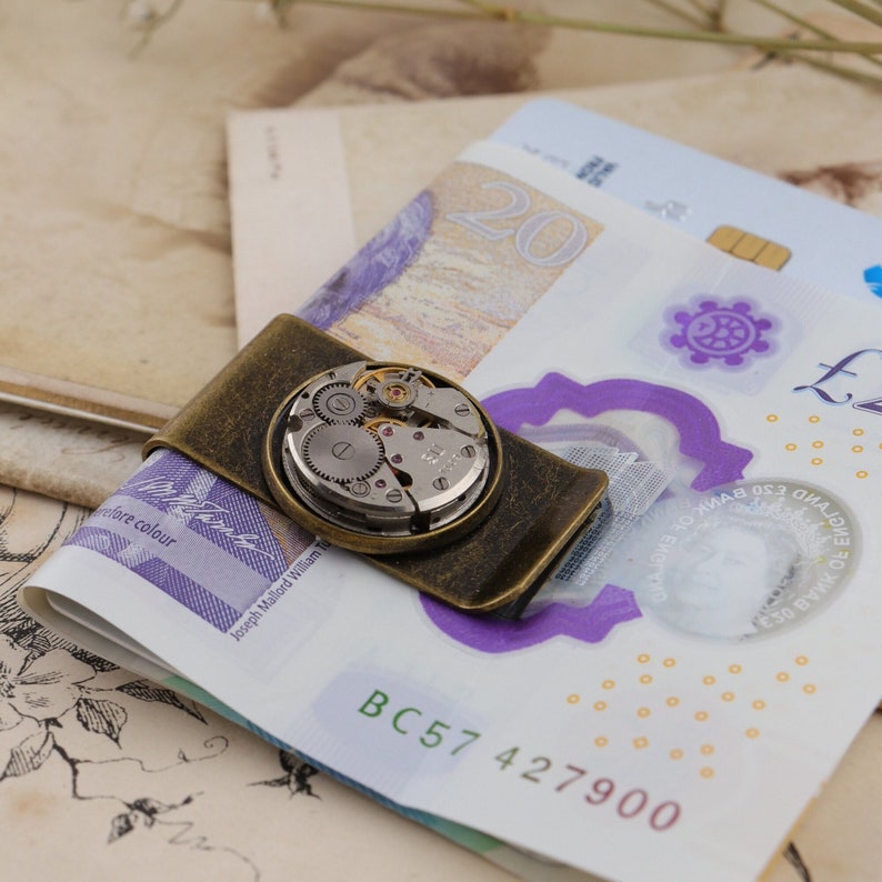 May include: A vintage-style money clip in antique brass finish, featuring a watch mechanism. The clip holds a stack of British twenty-pound notes. The watch mechanism is round and has visible gears and other internal components.