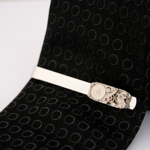 Puede incluir: Un pasador de corbata plateado con un dise&ntilde;o de movimiento de reloj. El pasador de corbata est&aacute; en una corbata negra con un patr&oacute;n de c&iacute;rculo repetido.