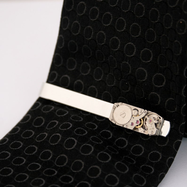 Puede incluir: Un pasador de corbata plateado con un dise&ntilde;o de movimiento de reloj. El pasador de corbata est&aacute; en una corbata negra con un patr&oacute;n de c&iacute;rculo repetido.