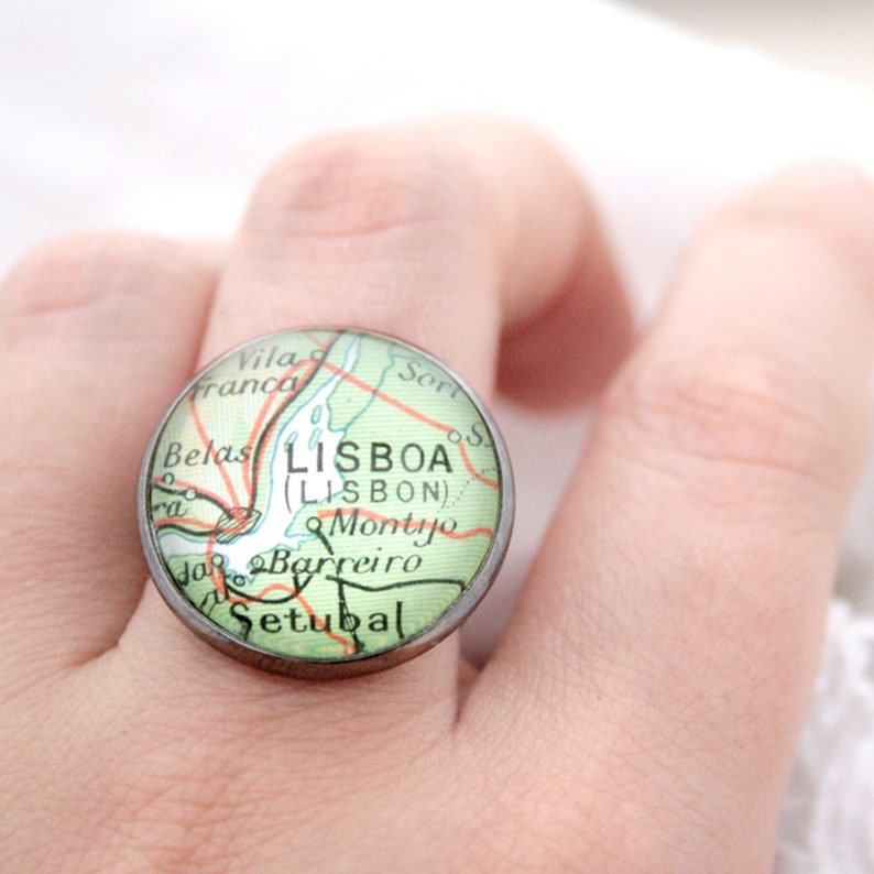 May include: A silver ring with a round glass cabochon featuring a vintage map of Lisbon, Portugal. The map shows the city's name in both Portuguese and English, "LISBOA (LISBON)".