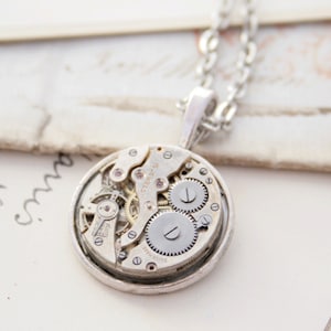 May include: Silver necklace with a round pendant featuring a vintage watch movement. The pendant is open and shows the intricate gears and springs of the watch mechanism.