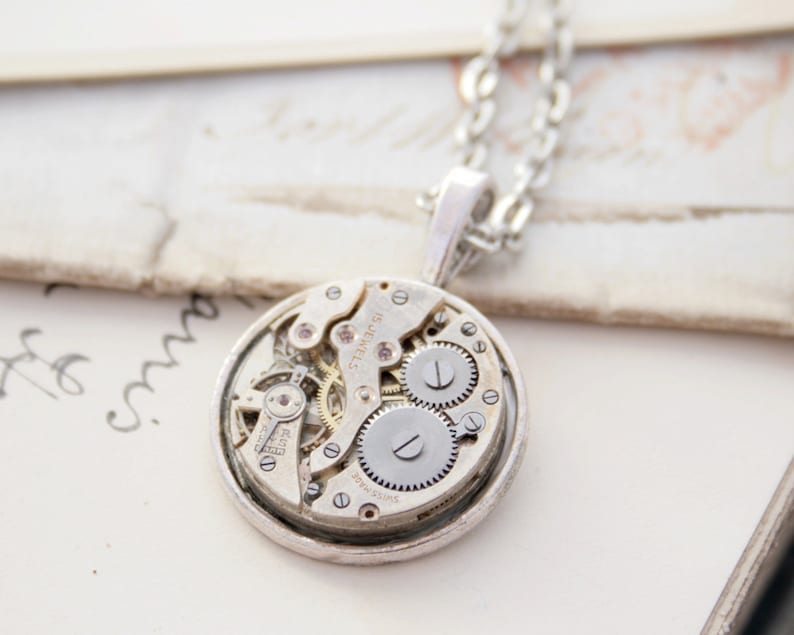 May include: Silver necklace with a round pendant featuring a vintage watch movement. The pendant is open and shows the intricate gears and springs of the watch mechanism.