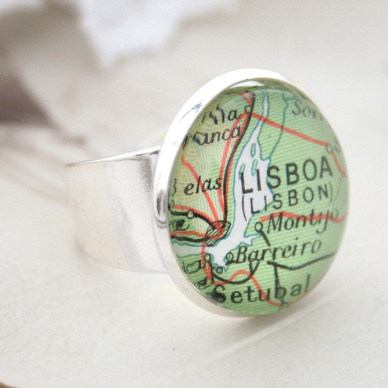 May include: A silver ring with a round glass cabochon featuring a vintage map of Lisbon, Portugal. The map shows the city's name in both Portuguese and English, "LISBOA (LISBON)", along with surrounding towns and geographical features.