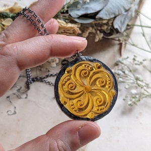 Puede incluir: Un colgante de vidrio dorado con un dise&ntilde;o floral detallado, enmarcado en metal negro, colgado de una cadena plateada.