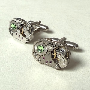 May include: A pair of silver-toned cufflinks featuring exposed mechanical watch movements. Each piece displays intricate metallic gears, small gold-colored wheels, and a single faceted light green stone set within the complex clockwork assembly.