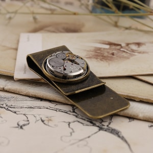 May include: A vintage-style money clip with a brass finish. The clip features a circular inset showcasing the intricate gears and components of a watch movement. The design combines functionality with a steampunk aesthetic.
