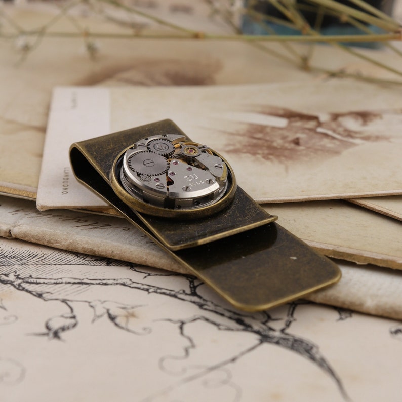 May include: A vintage-style money clip with a brass finish. The clip features a circular inset showcasing the intricate gears and components of a watch movement. The design combines functionality with a steampunk aesthetic.
