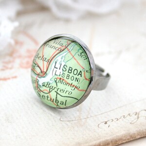 May include: A silver ring with a glass cabochon featuring a vintage map of Lisbon, Portugal. The map shows the city's name in both Portuguese and English, "LISBOA (LISBON)".