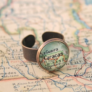 May include: A bronze ring with a glass cabochon featuring a vintage map of Venezuela with the city of Caracas highlighted. The map is green, brown, and yellow.
