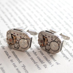 May include: A pair of silver cufflinks with a mechanical watch movement design. The cufflinks feature visible gears, tiny red jewels, and intricate metalwork, set against a blurred background of printed text.