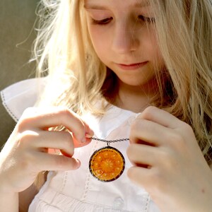 Puede incluir: Una ni&ntilde;a peque&ntilde;a con un vestido blanco sostiene un collar con un gran colgante redondo de color naranja. El colgante tiene un dise&ntilde;o floral detallado.