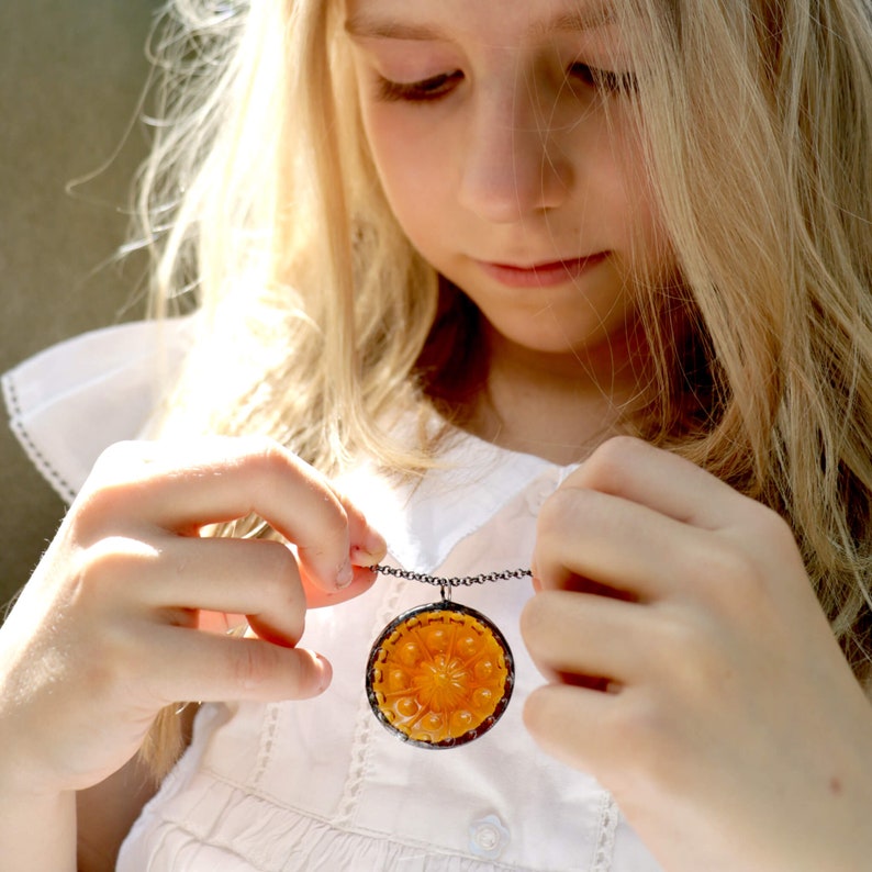 Puede incluir: Una ni&ntilde;a peque&ntilde;a con un vestido blanco sostiene un collar con un gran colgante redondo de color naranja. El colgante tiene un dise&ntilde;o floral detallado.