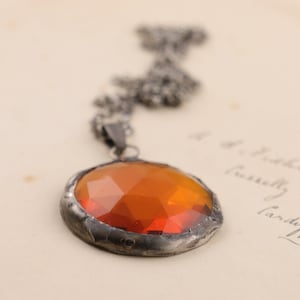 Puede incluir: Un collar de cadena de plata con un colgante redondo de vidrio naranja. El colgante est&aacute; engastado en un marco de plata y tiene un dise&ntilde;o facetado.