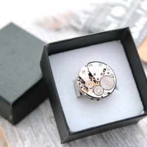 May include: A silver ring with a circular watch face design. The watch face is made of metal and has visible gears and a small ruby-colored jewel.