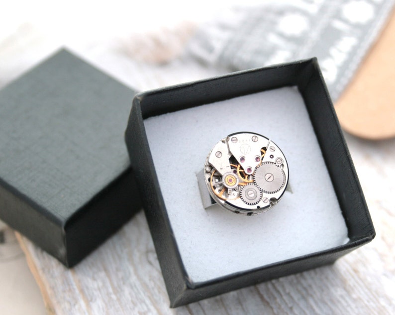 May include: A silver ring with a circular watch face design. The watch face is made of metal and has visible gears and a small ruby-colored jewel.