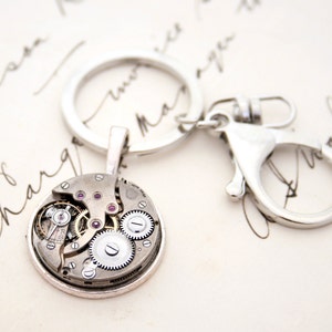 May include: A silver key chain with a round, silver charm featuring a visible clockwork mechanism with pink jewels. The charm is attached to a silver key ring and a silver lobster clasp.