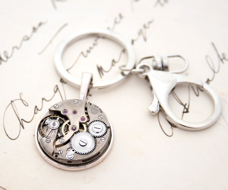 May include: A silver key chain with a round, silver charm featuring a visible clockwork mechanism with pink jewels. The charm is attached to a silver key ring and a silver lobster clasp.