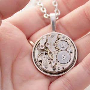 May include: Silver necklace with a round pendant featuring a visible clockwork mechanism. The pendant reads "Swiss Made" and "15 Jewels".