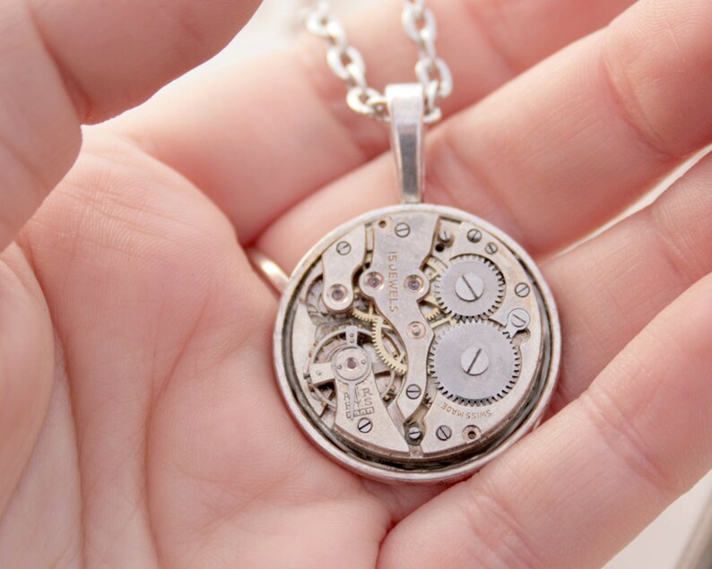 May include: Silver necklace with a round pendant featuring a visible clockwork mechanism. The pendant reads "Swiss Made" and "15 Jewels".